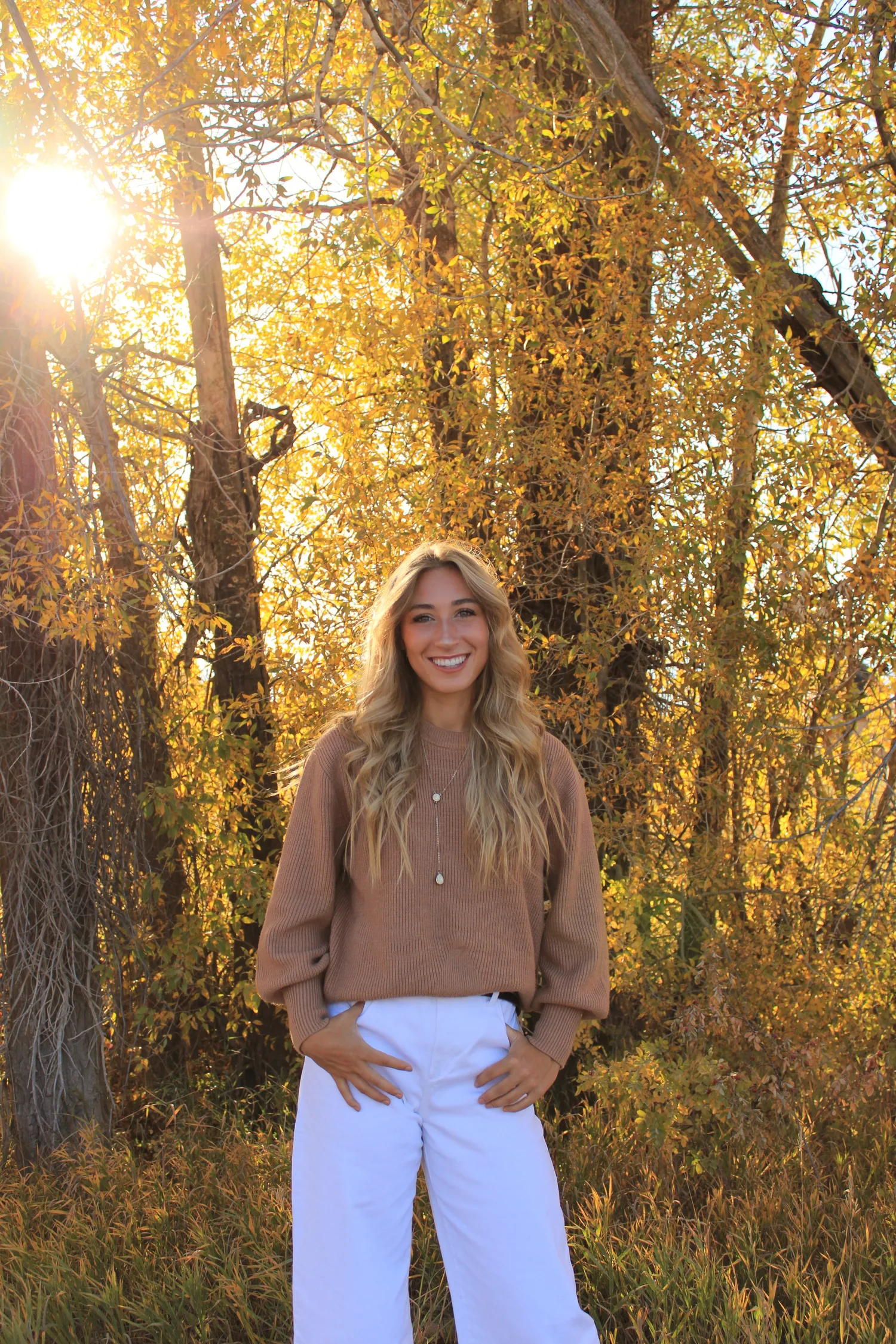 Senior portrait of a young woman in a tan sweater among glowing yellow fall trees