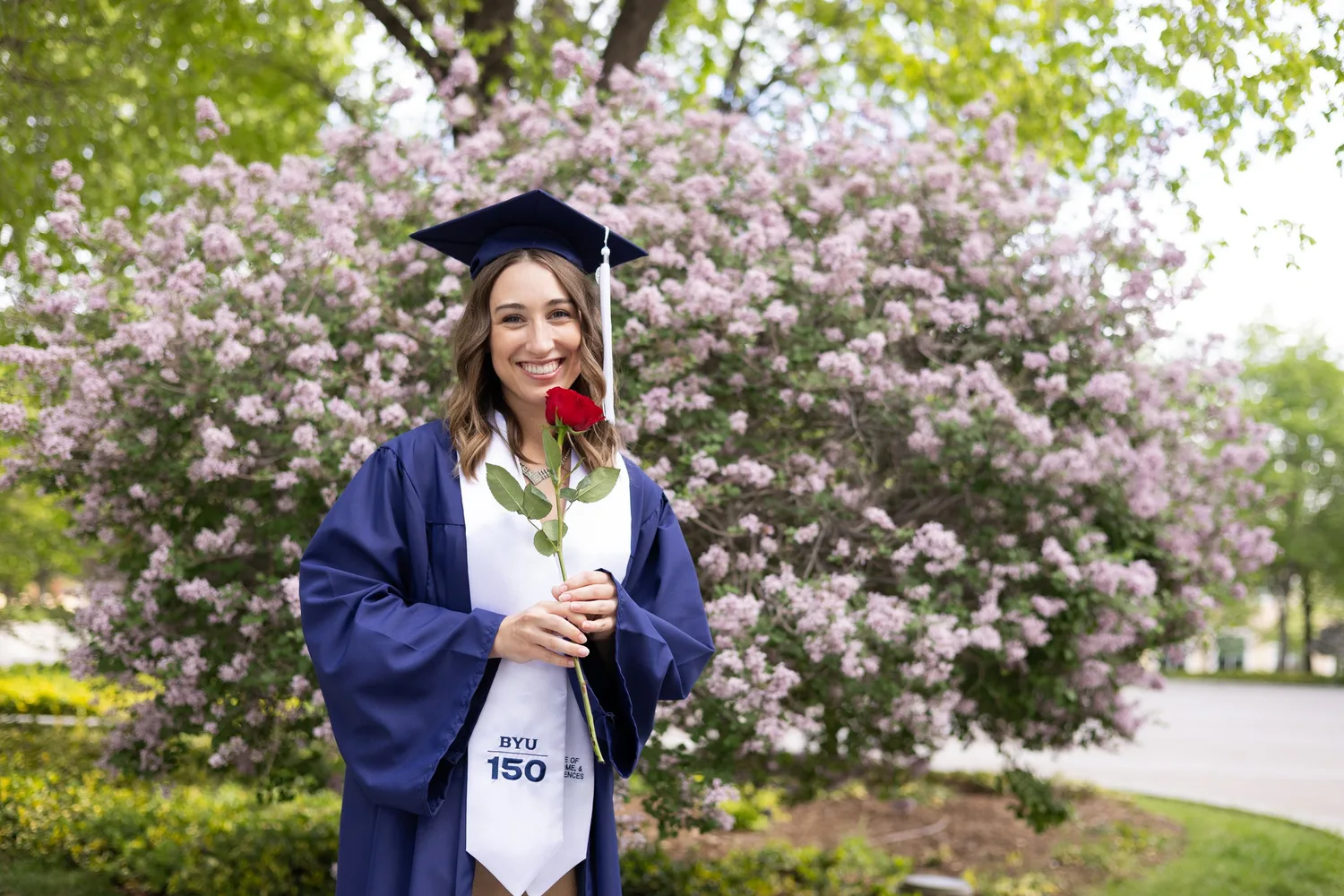 BYU graduate in a navy gown holding a red rose in front of a flowering lilac