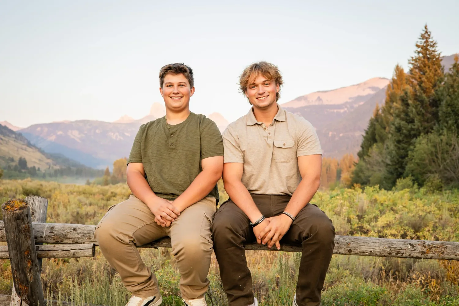 Two young men sitting on a wooden fence with a mountain valley behind them