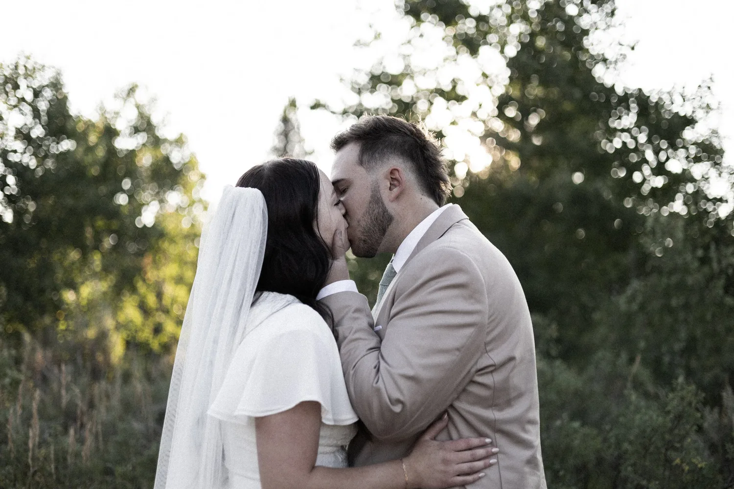 Bride and groom kissing at sunset with trees behind them