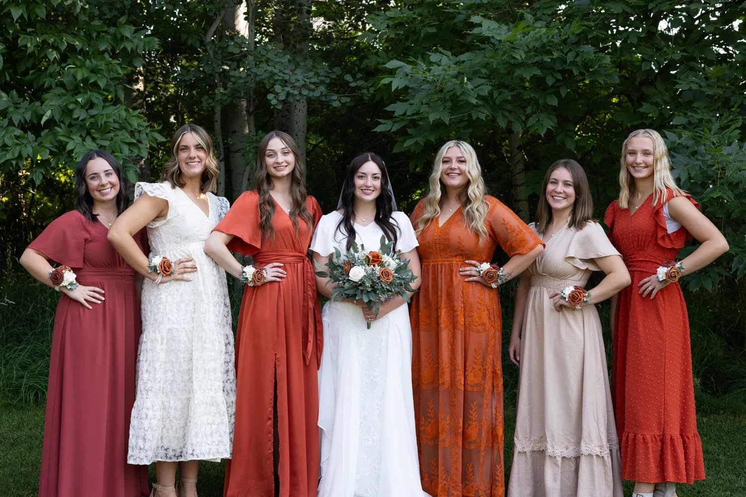 Bride with bridesmaids in rust, terracotta and blush dresses in front of green trees