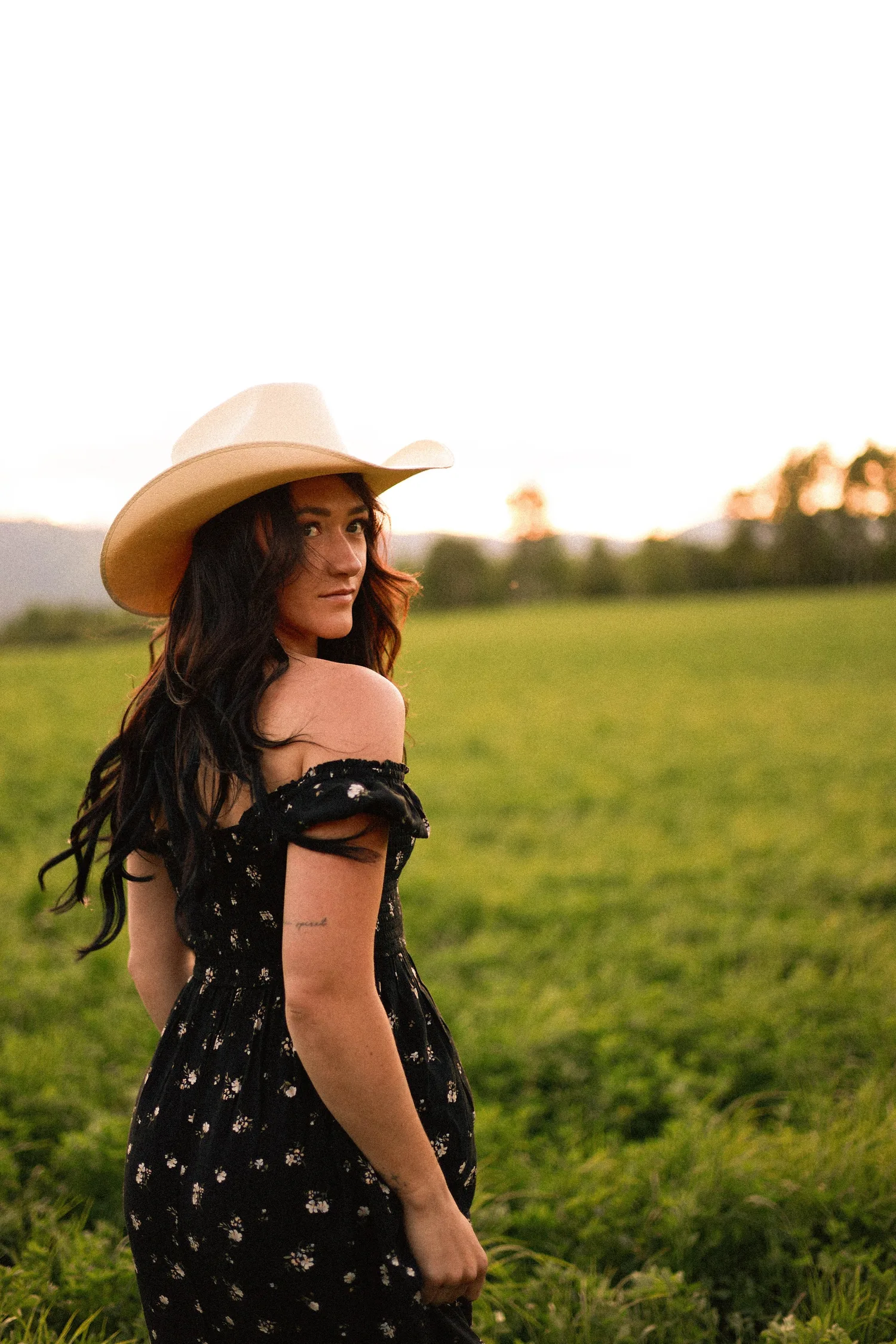 Young woman in a cowboy hat and black floral dress looking over her shoulder in a green field