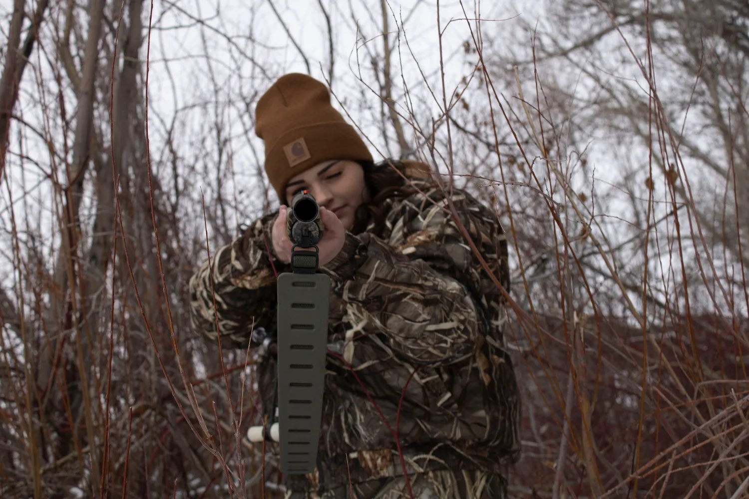 Hunter in camouflage and a beanie aiming a shotgun from the brush