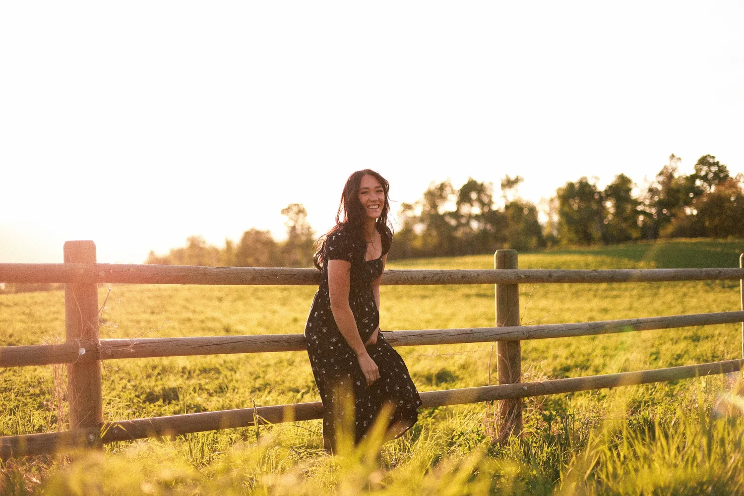 Woman in a black floral dress smiling by a wooden fence in a golden-hour field