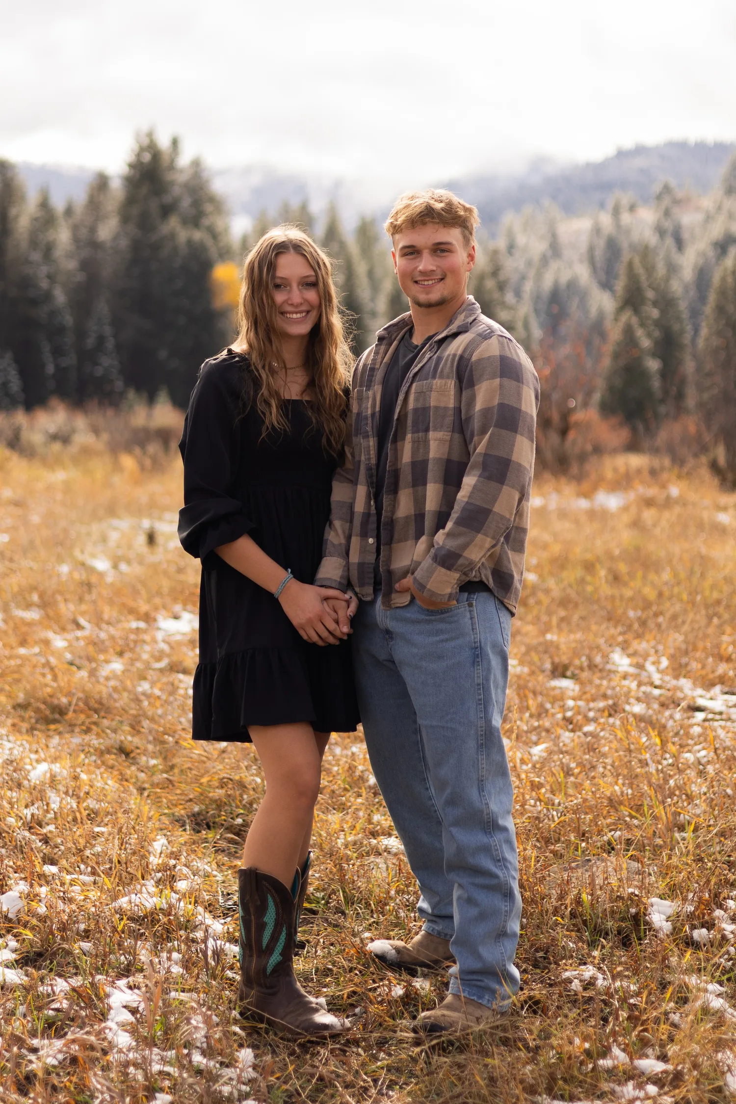 Couple in flannel and cowboy boots standing in a snow-dusted autumn meadow