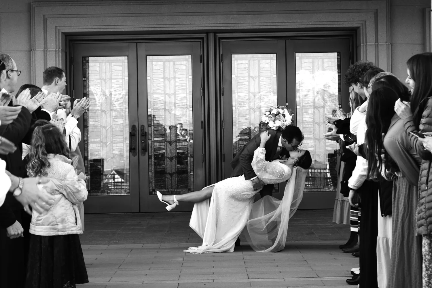 Black-and-white photo of a bride and groom kissing as guests cheer outside the temple doors