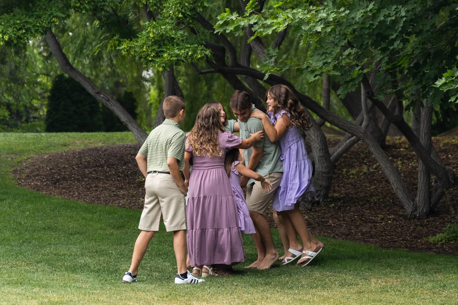 Siblings hugging and laughing on a lawn under shady trees