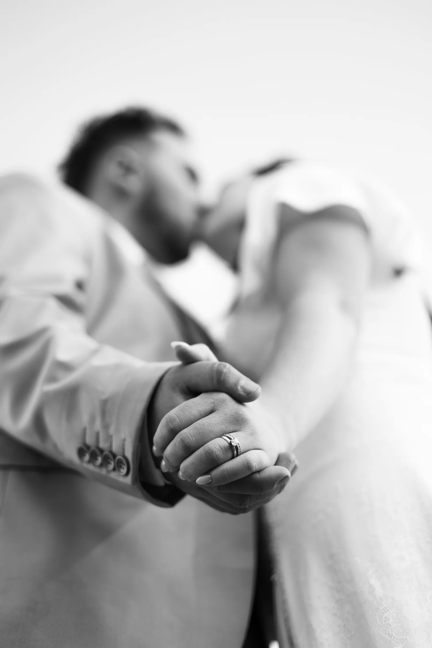 Black-and-white close-up of a couple kissing while holding hands, showing the ring