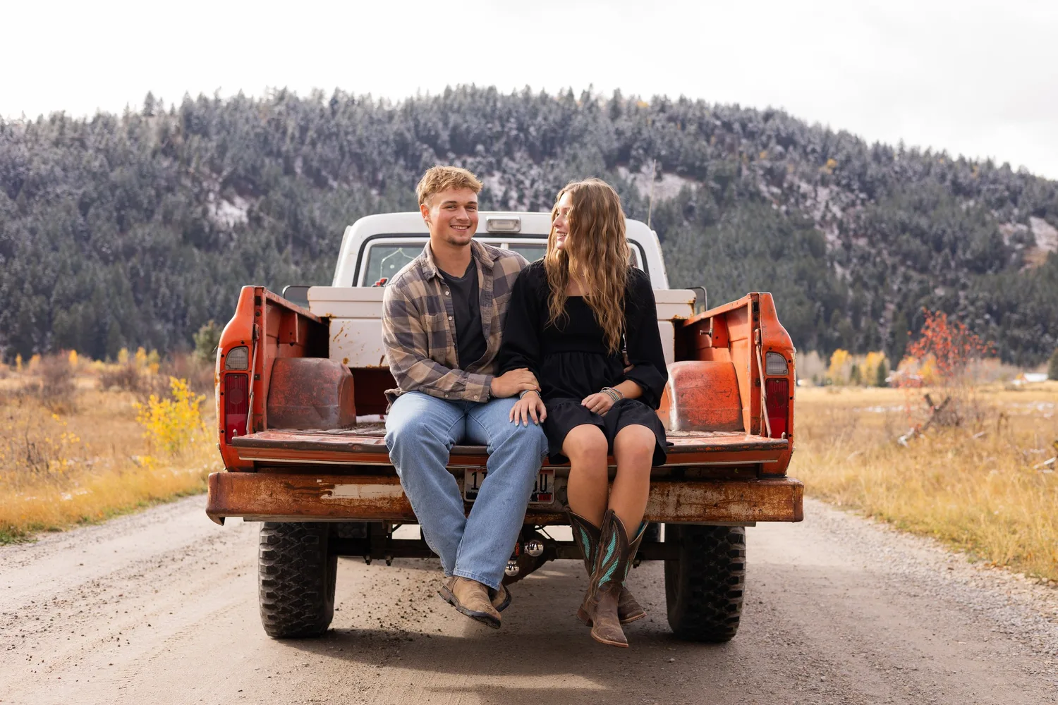 Couple sitting on an orange vintage truck tailgate on a dirt road below snowy hills