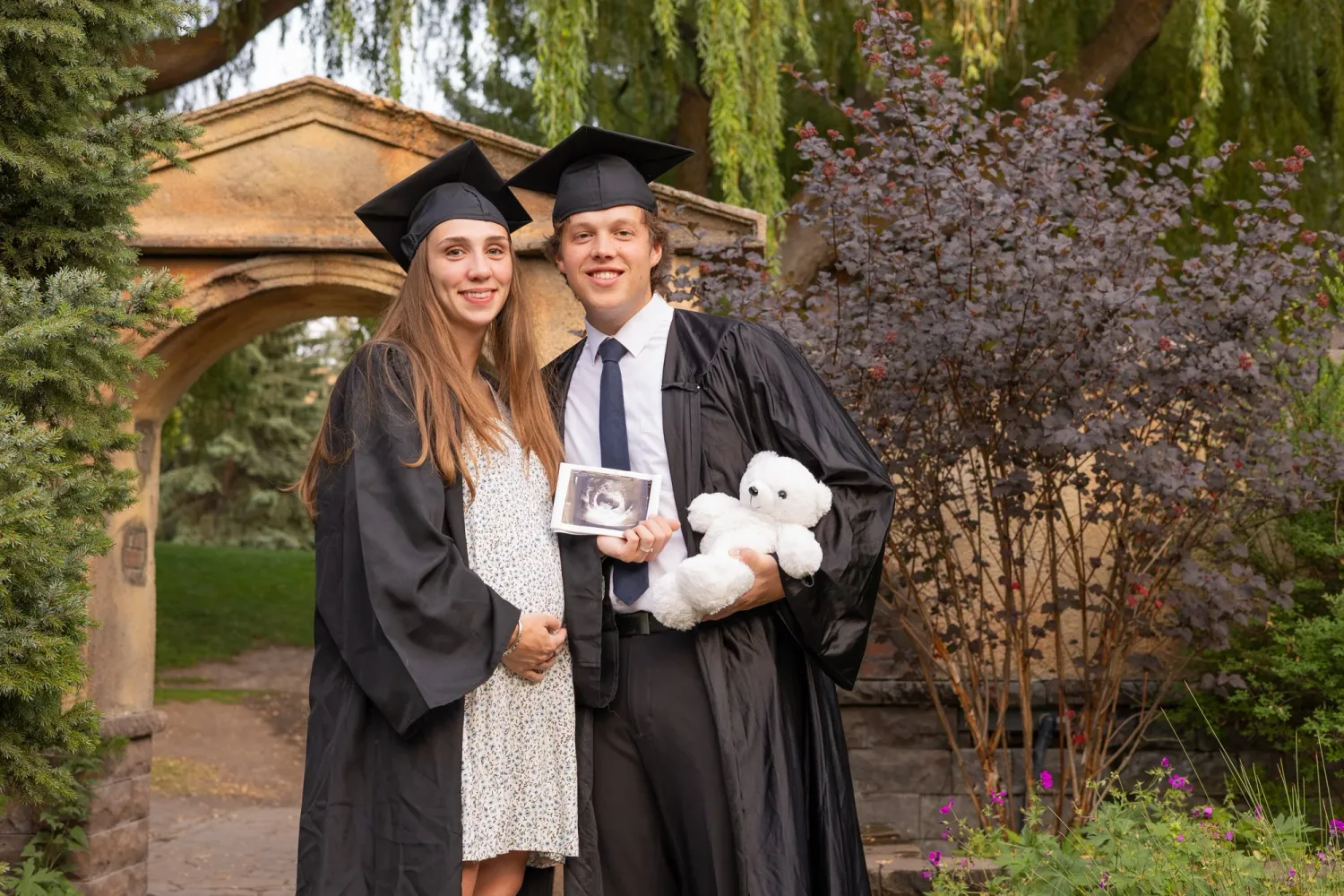 Graduating couple in caps and gowns holding an ultrasound photo and a teddy bear for a pregnancy announcement