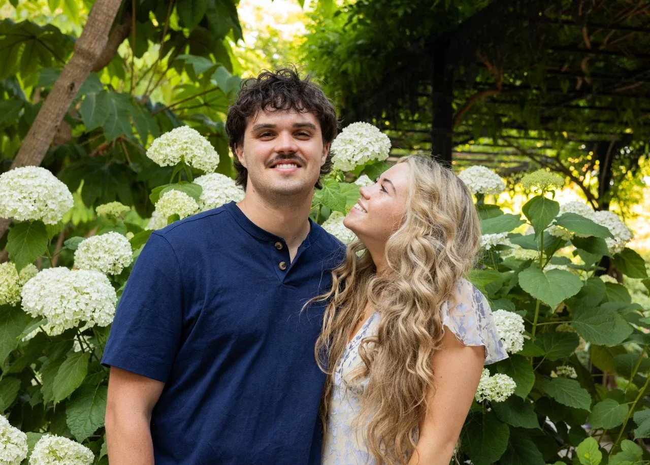 Engaged couple smiling in front of blooming white hydrangeas