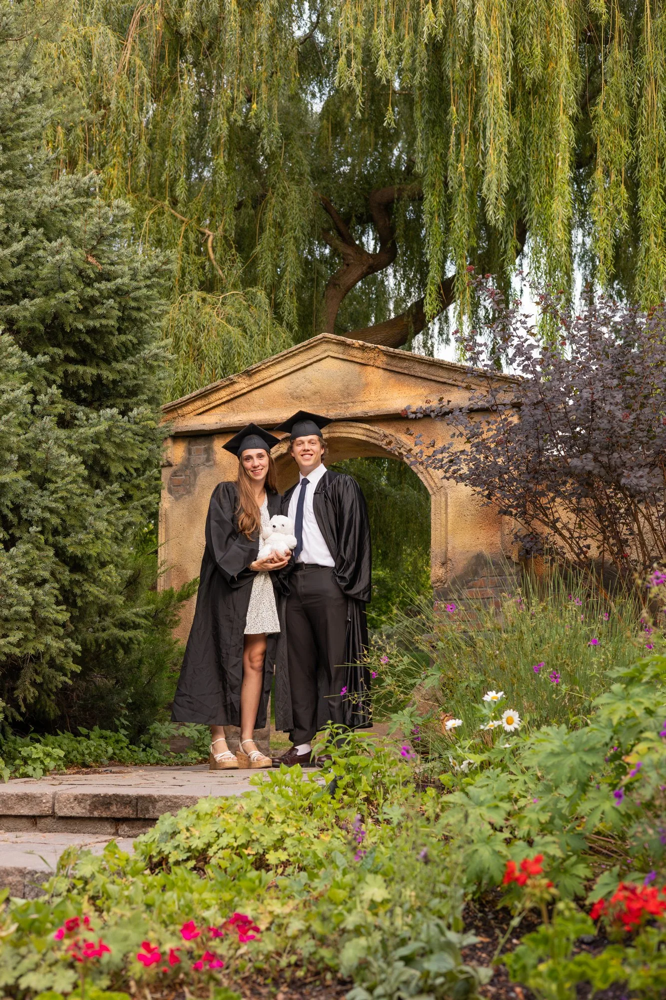 Graduating couple in caps and gowns under a stone arch in a flower garden