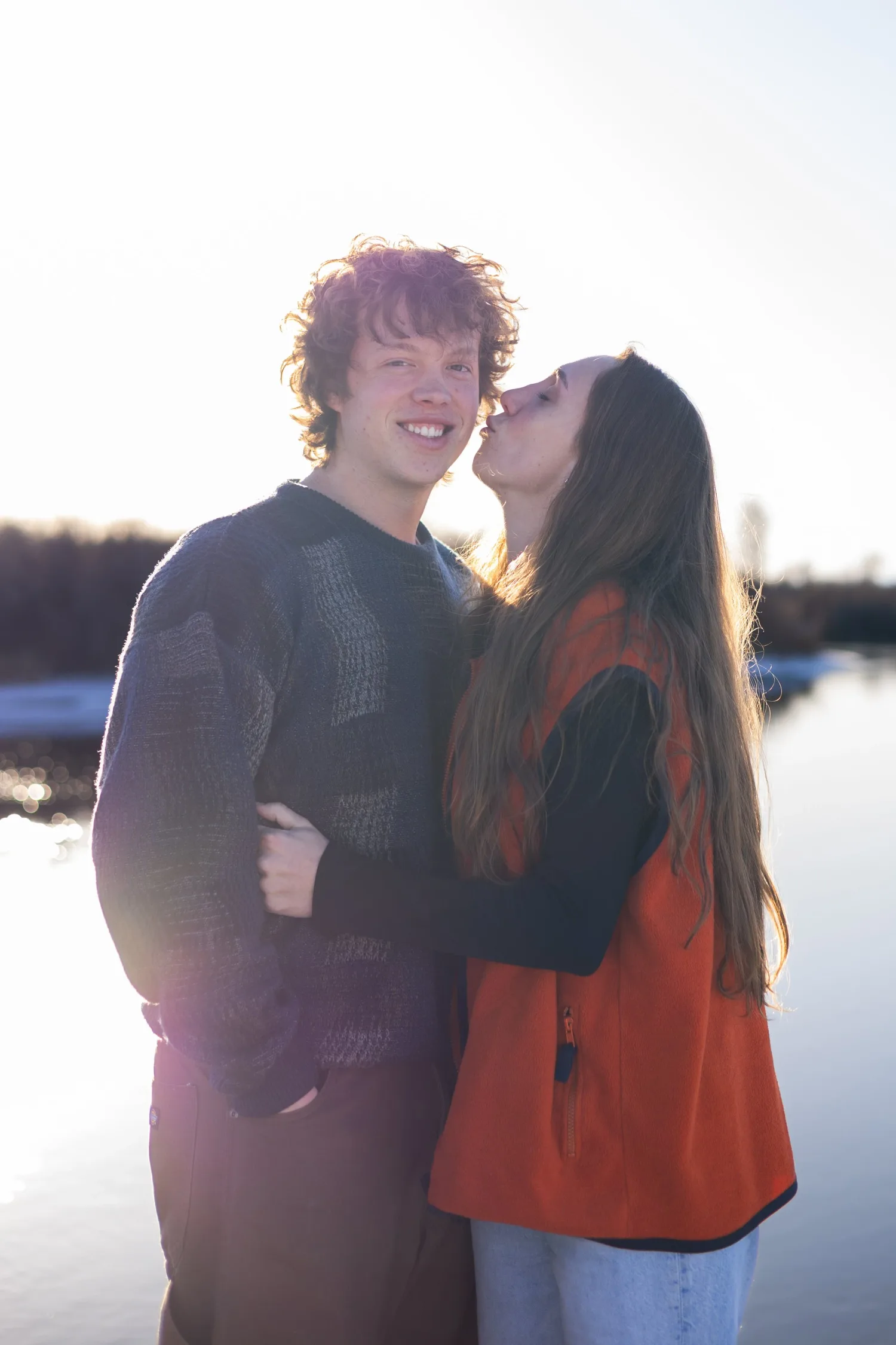Young woman kissing her boyfriend's cheek beside a lake at sunset