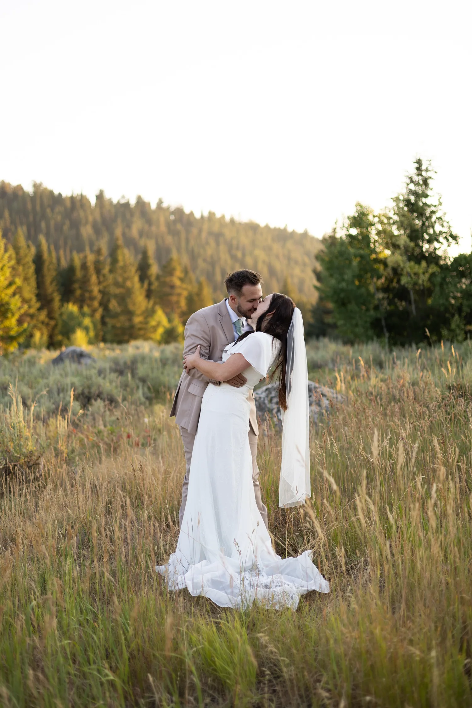 Bride and groom embracing in a mountain meadow with her veil blowing