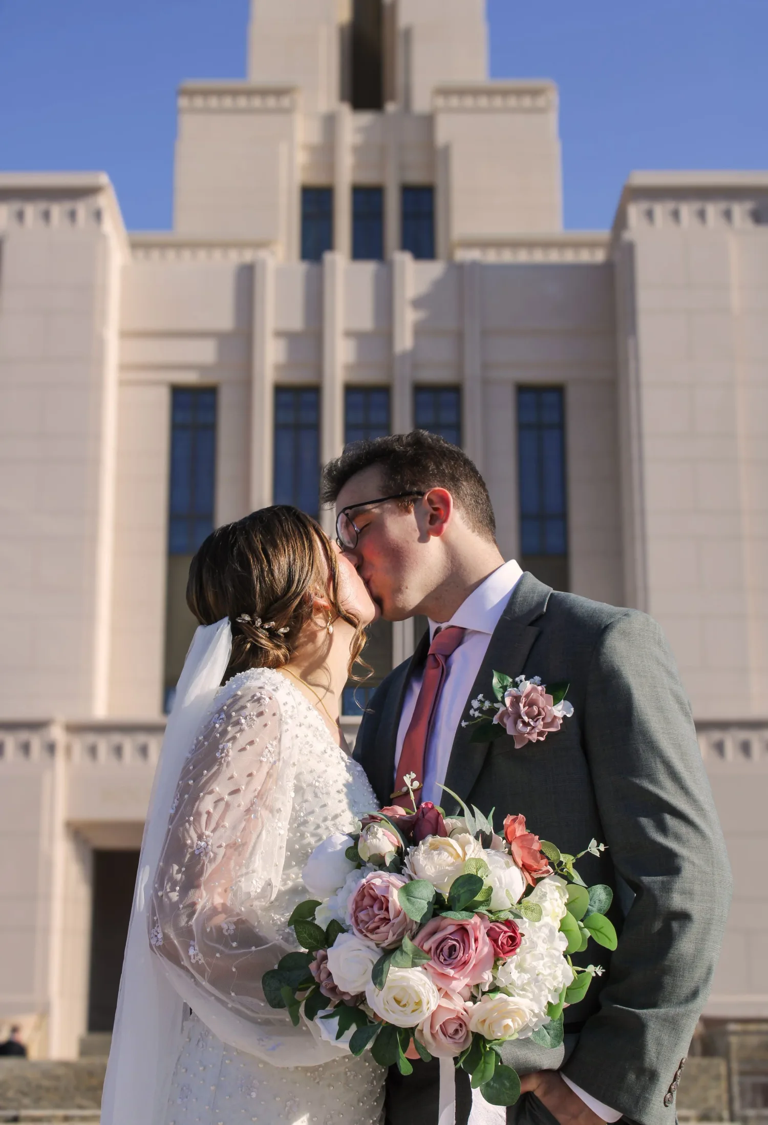 Bride and groom kissing in front of a white temple, bride holding a pink and white rose bouquet