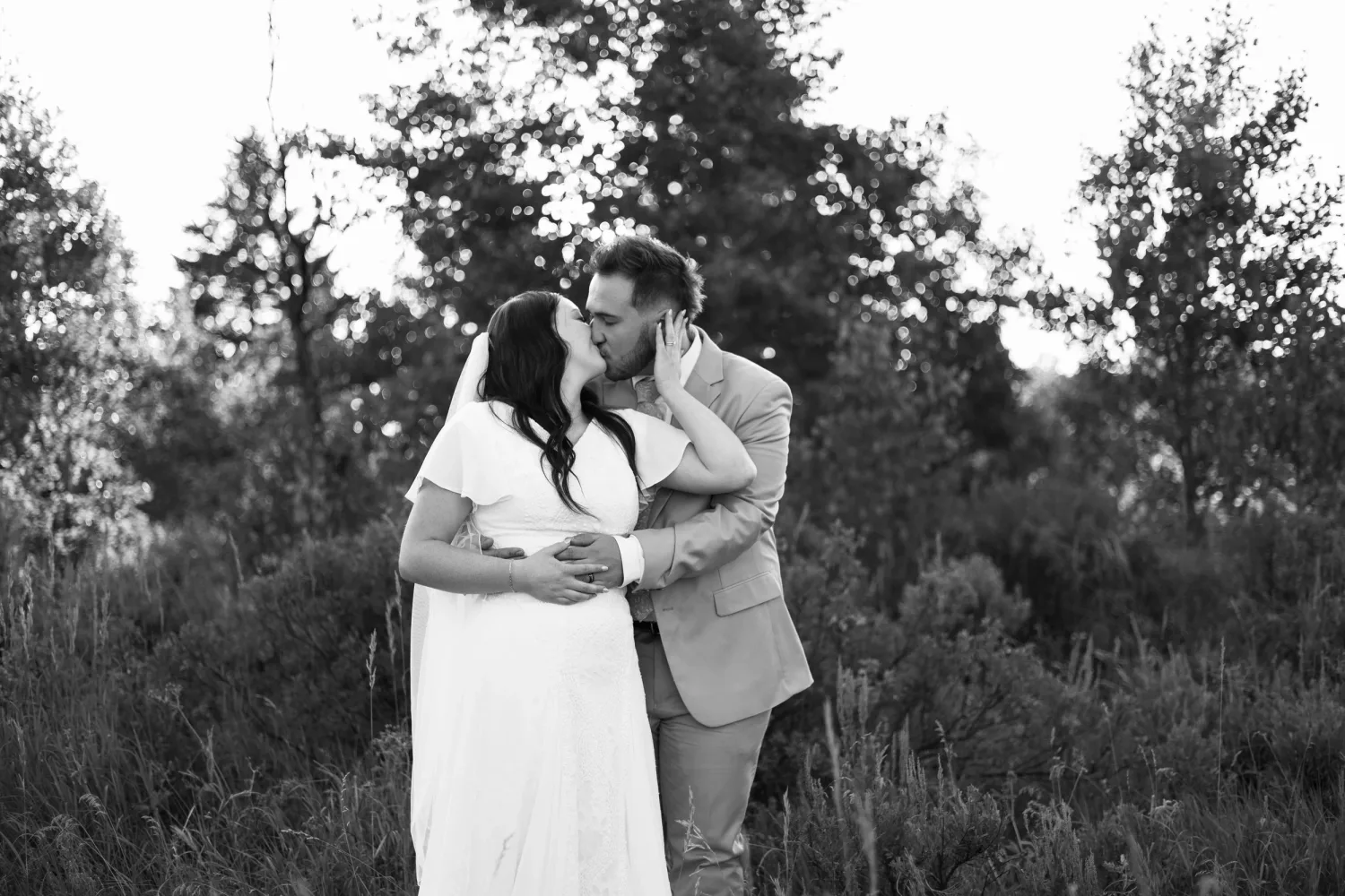 Black-and-white photo of a groom kissing his bride's forehead among trees