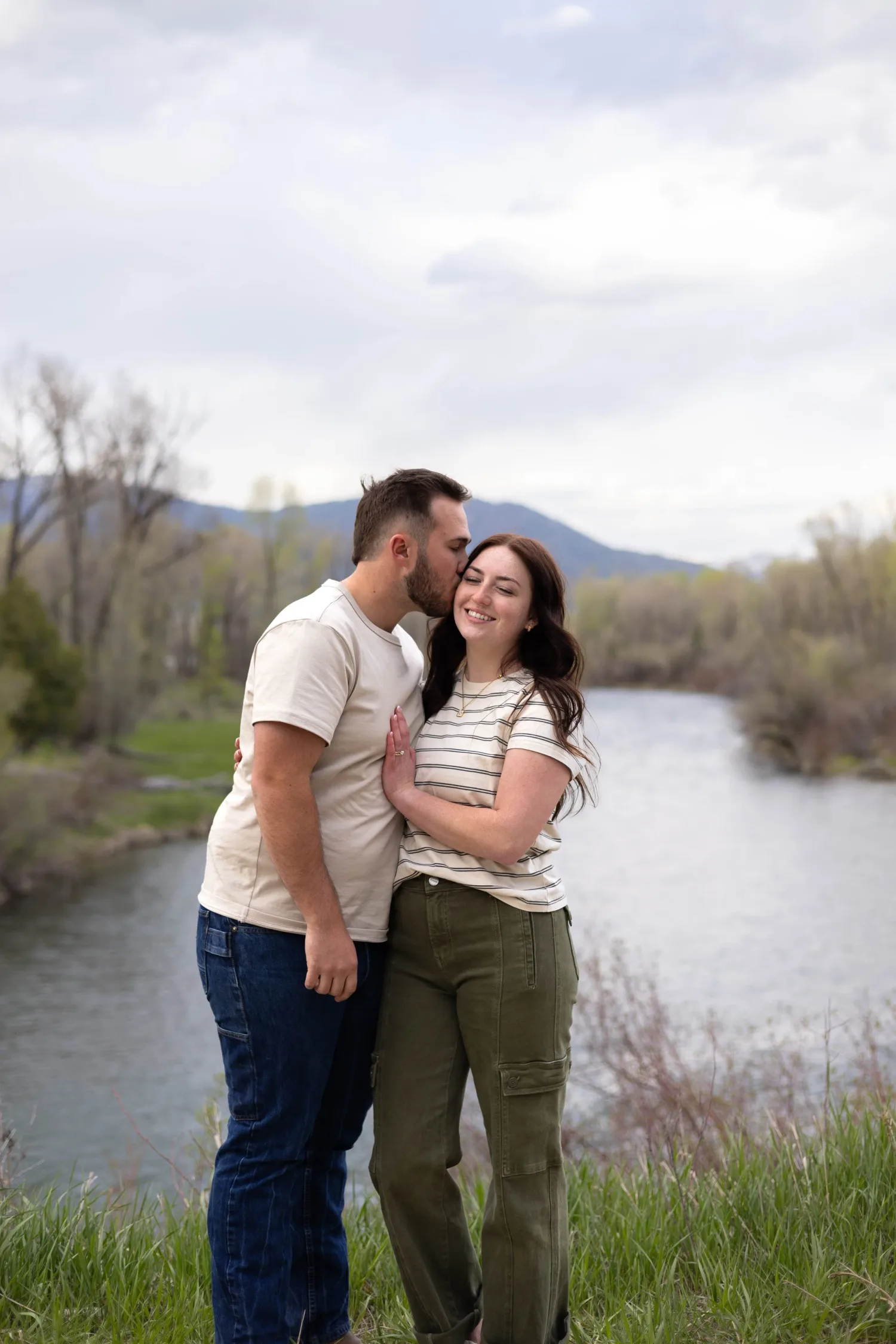 Man kissing his fiancée's head beside a river with mountains in the distance