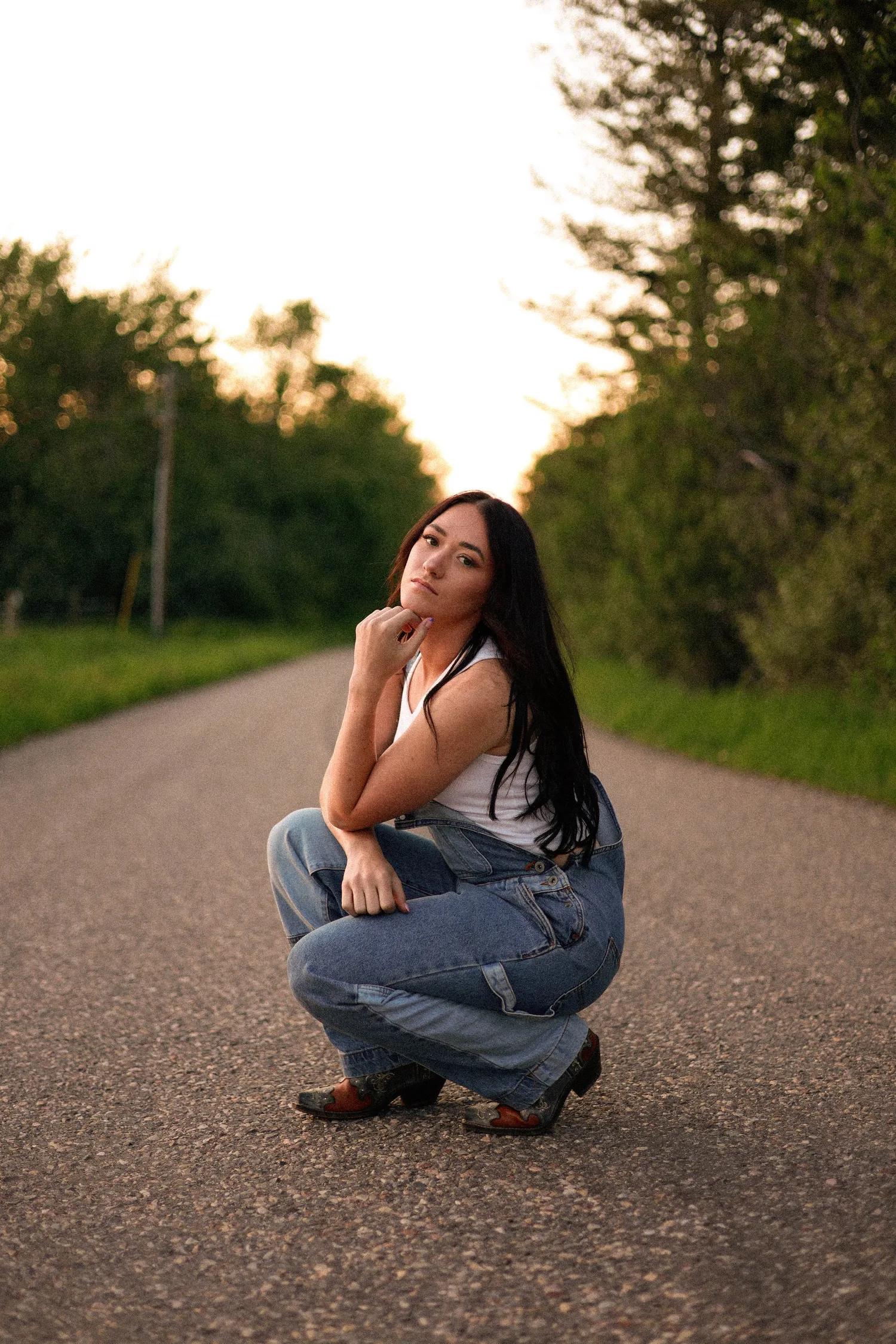 Senior portrait of a young woman crouching on a tree-lined road at sunset