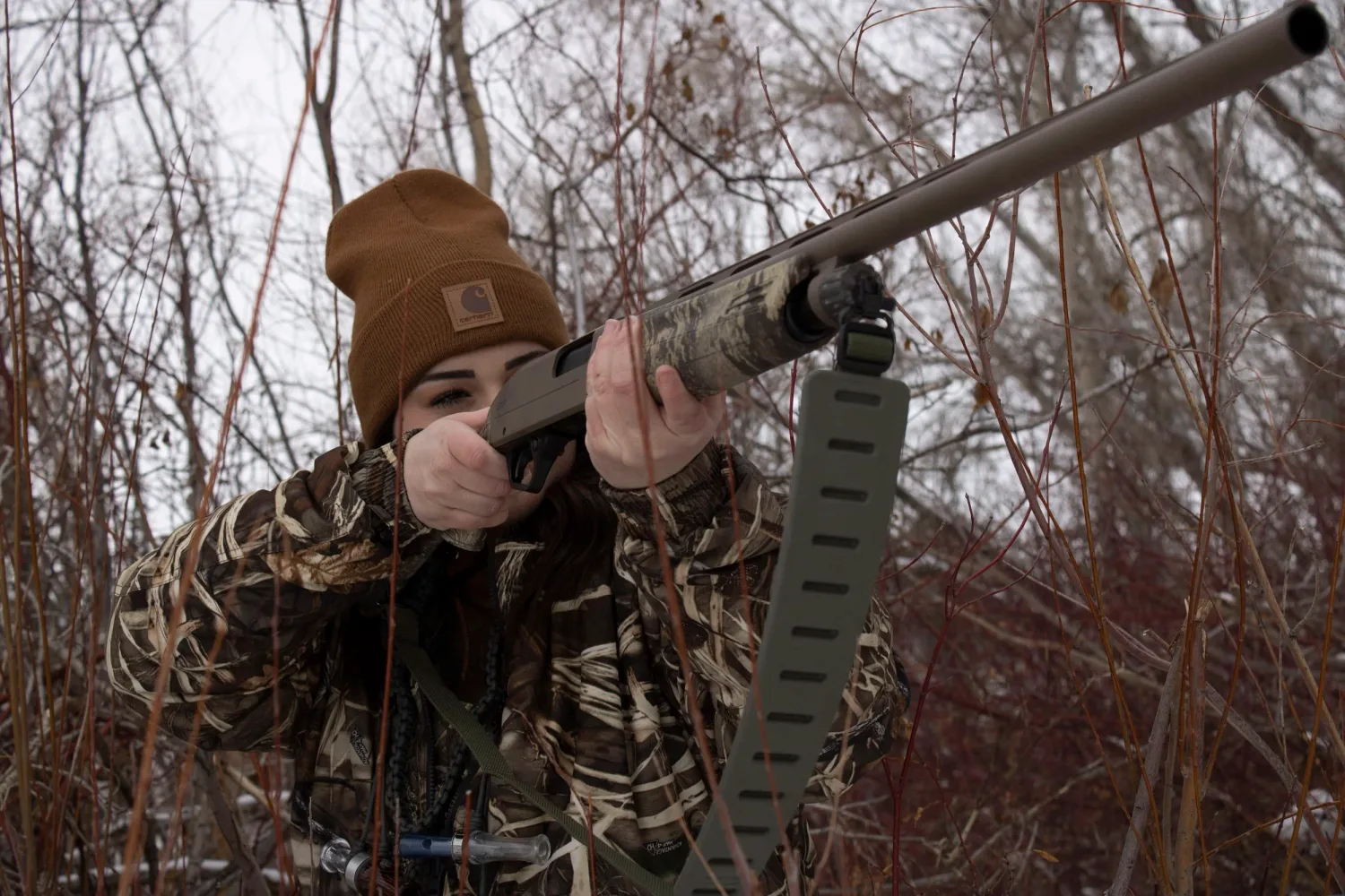 Hunter in camouflage aiming a shotgun through bare winter brush