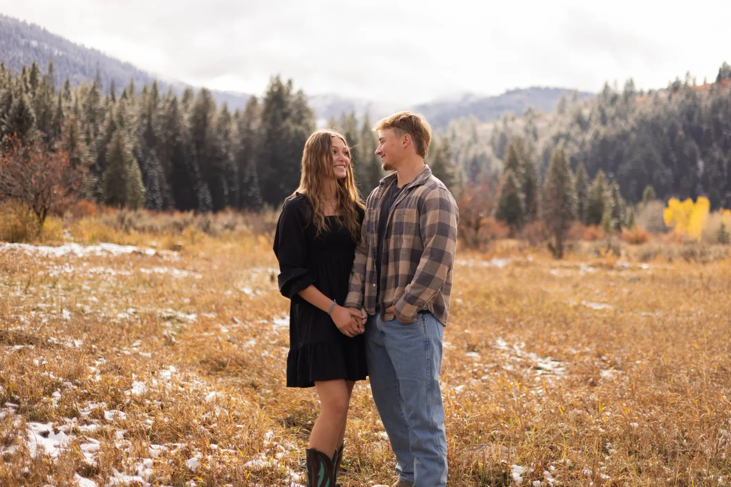 Couple holding each other in a snowy autumn meadow surrounded by pine-covered hills