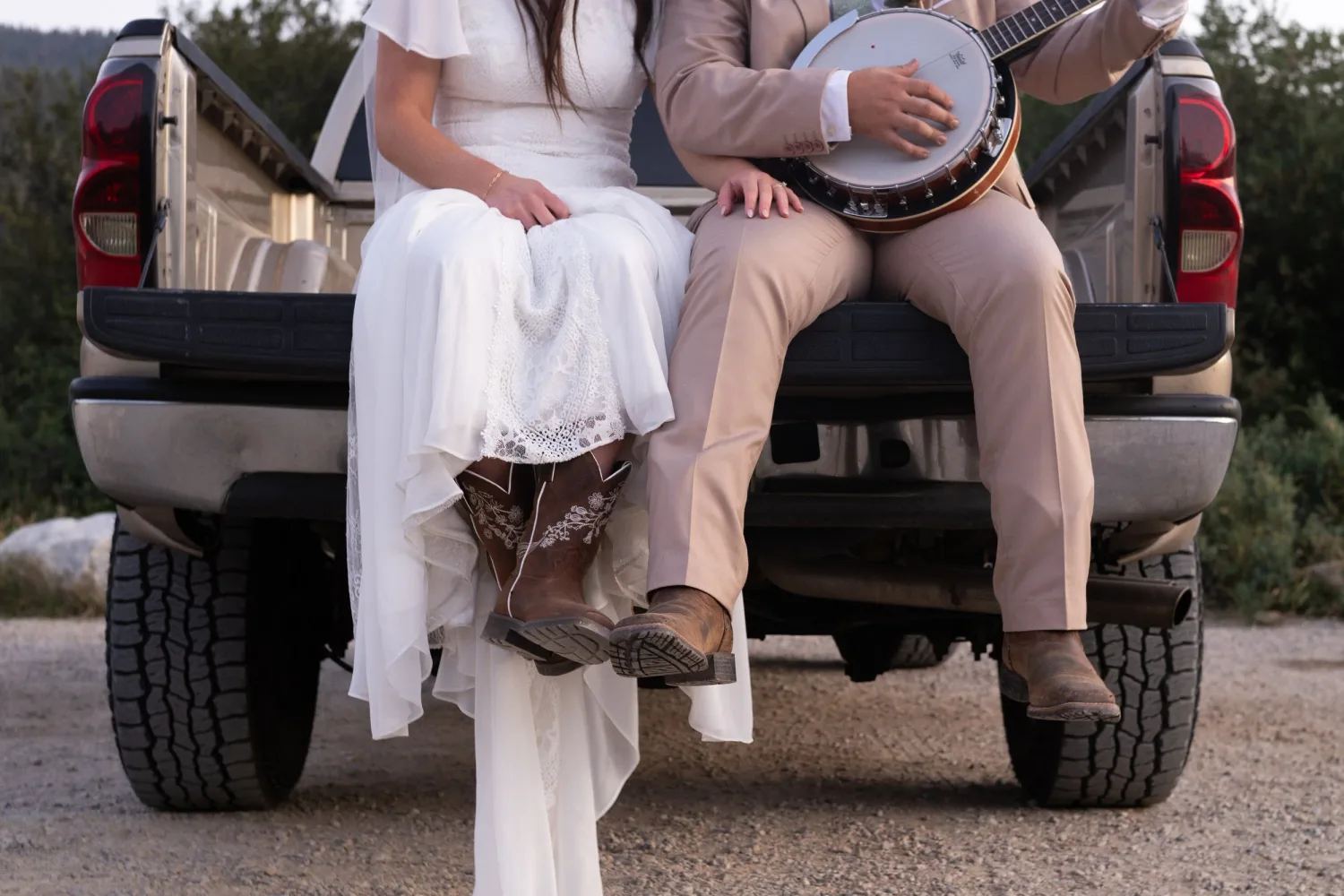 Bride in cowboy boots and groom with a banjo sitting on a pickup truck tailgate