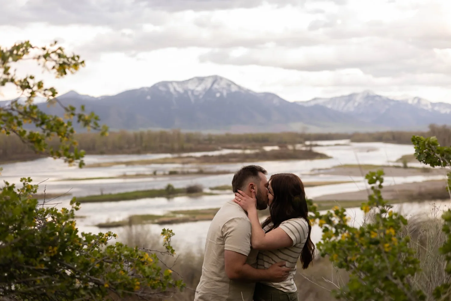Couple kissing on a bluff above a braided river with snow-capped mountains