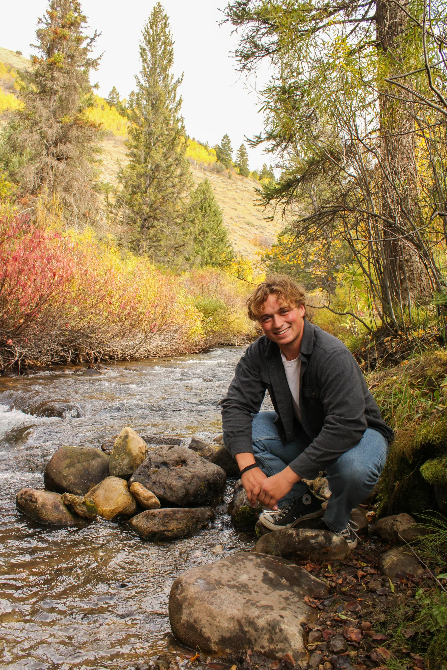 Senior portrait of a young man crouching by a rocky mountain stream in the fall