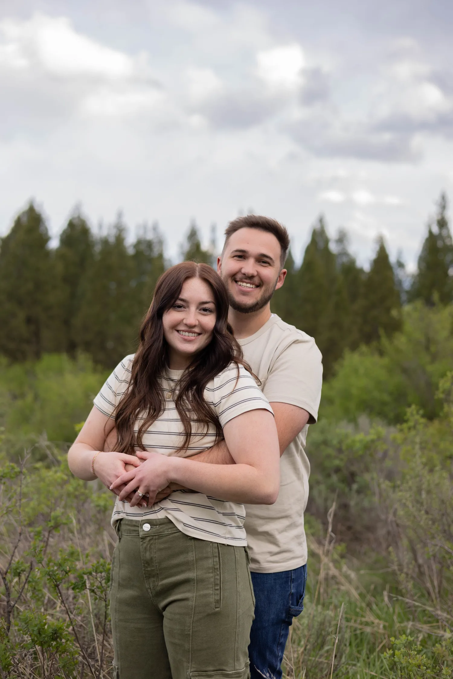 Smiling couple embracing in a green meadow lined with pine trees