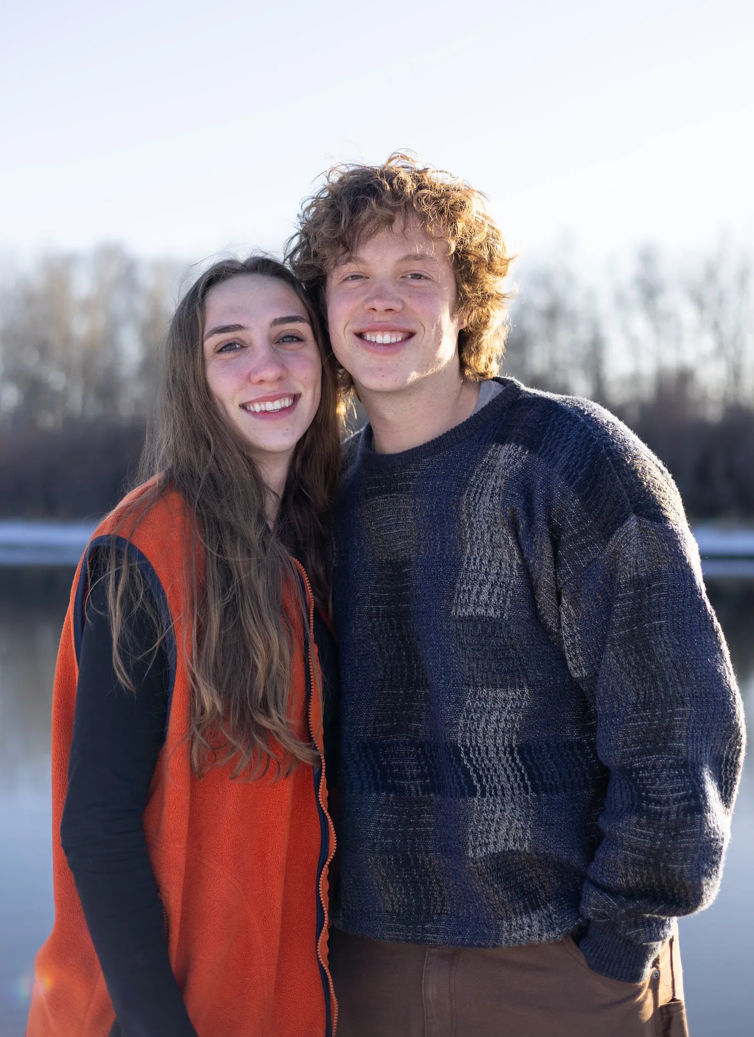Smiling couple in cozy sweaters by a river on a sunny winter day
