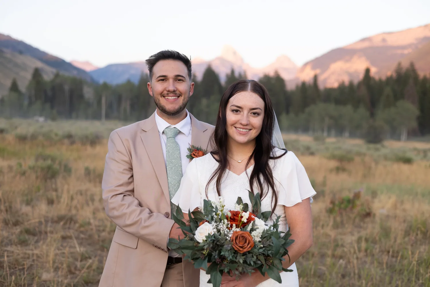 Bride and groom with a rust and white bouquet in a meadow with the Tetons at sunset