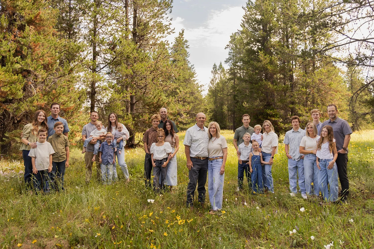 Large extended family portrait in a grassy meadow surrounded by pine trees