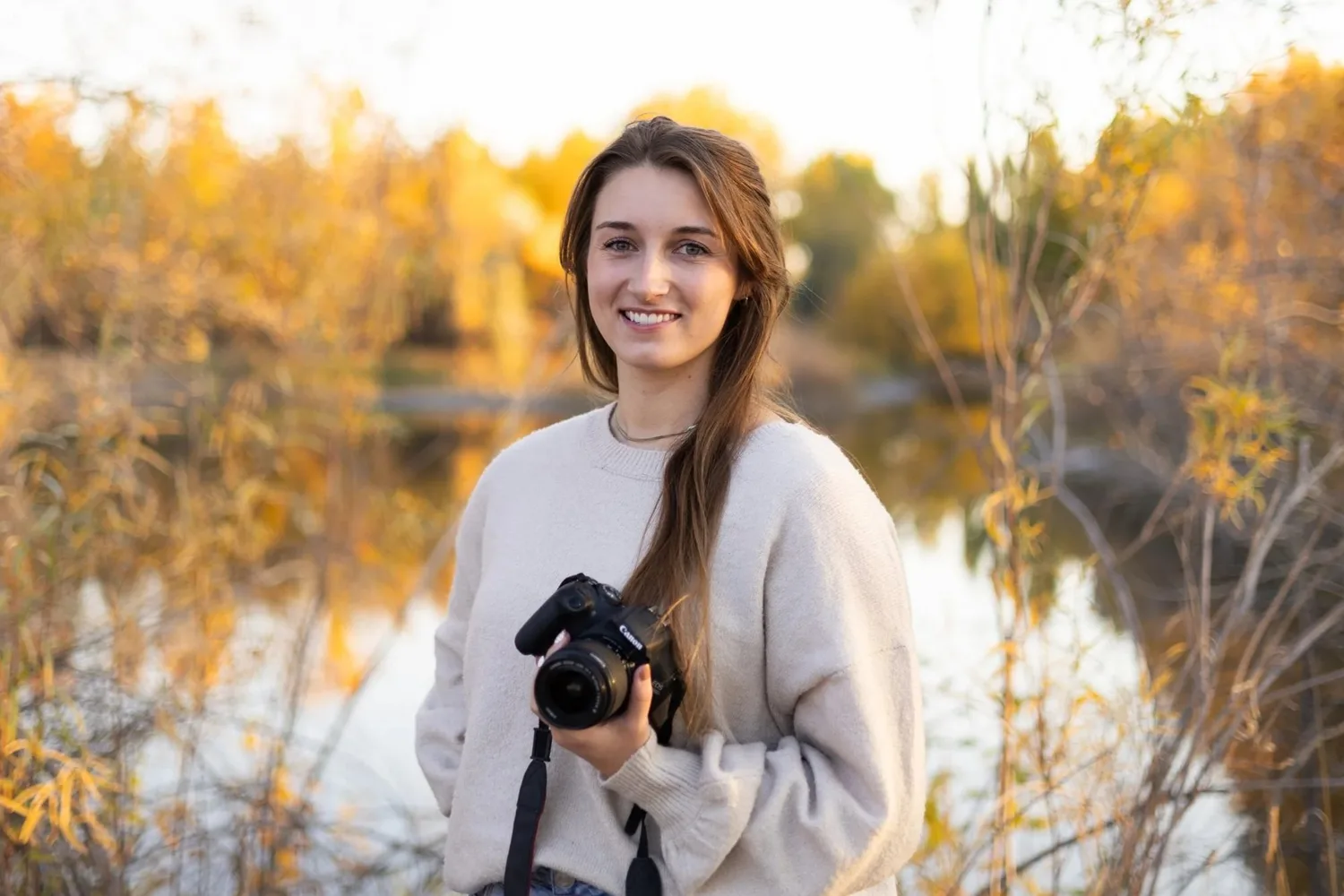 Toni Howell, photographer, holding her camera beside a pond with autumn trees