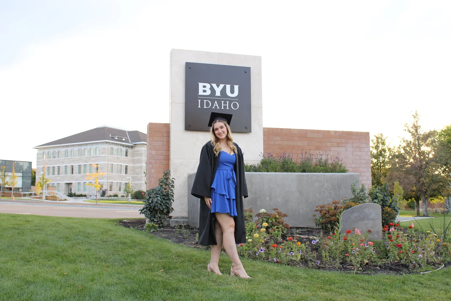 Graduate in cap and gown with a blue dress posing by the BYU-Idaho sign