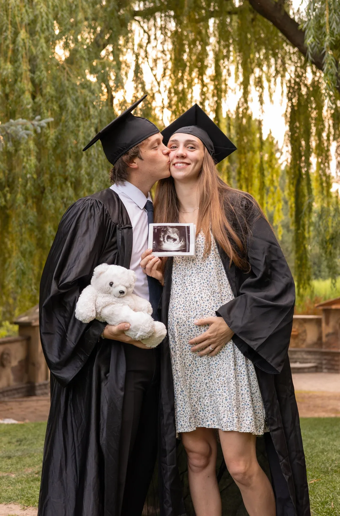 Graduating couple in caps and gowns kissing while holding an ultrasound photo and a teddy bear