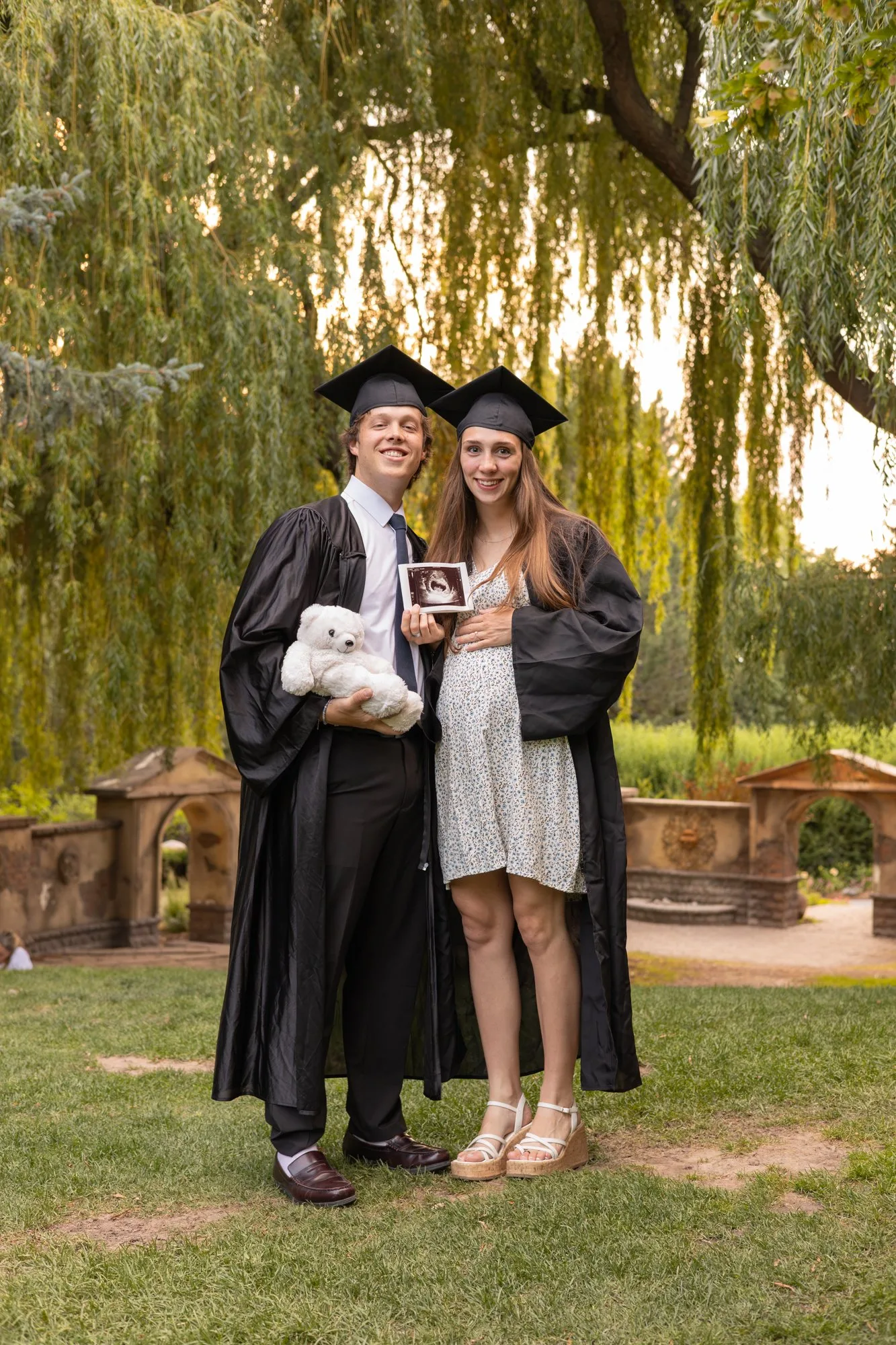 Graduating couple in caps and gowns under a willow tree holding an ultrasound and a teddy bear