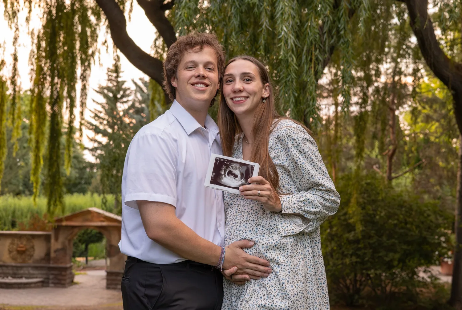 Expecting couple holding an ultrasound photo under willow trees