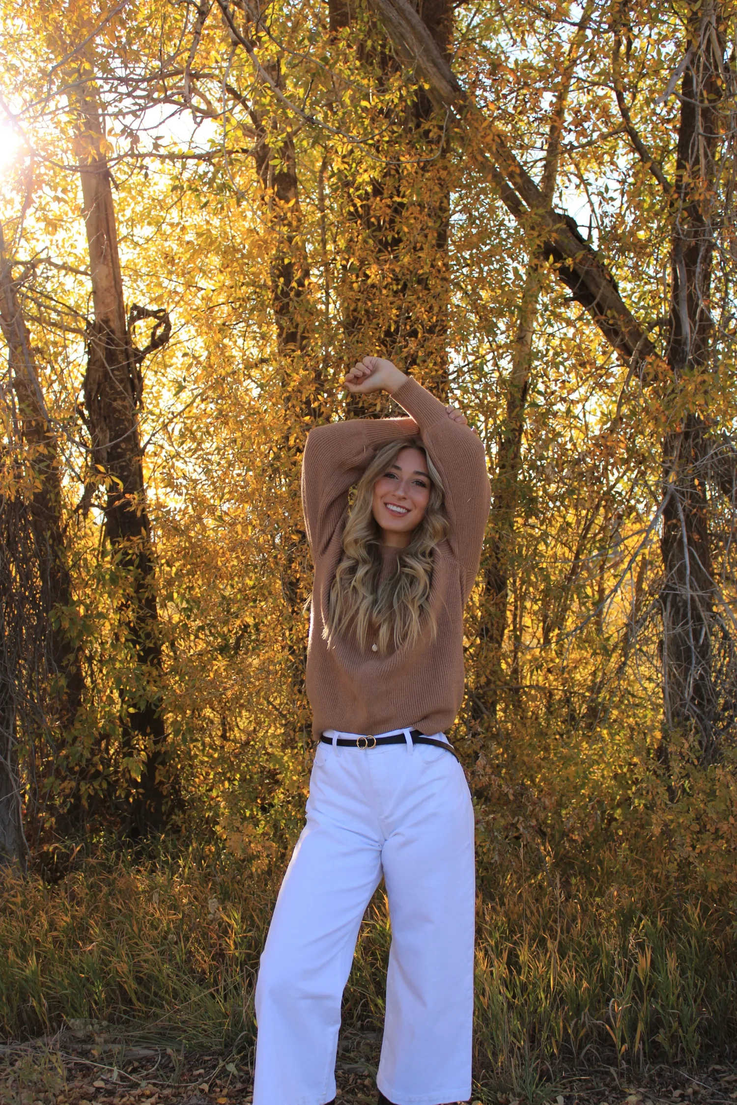 Senior portrait of a young woman in a tan sweater and white jeans in a golden autumn forest