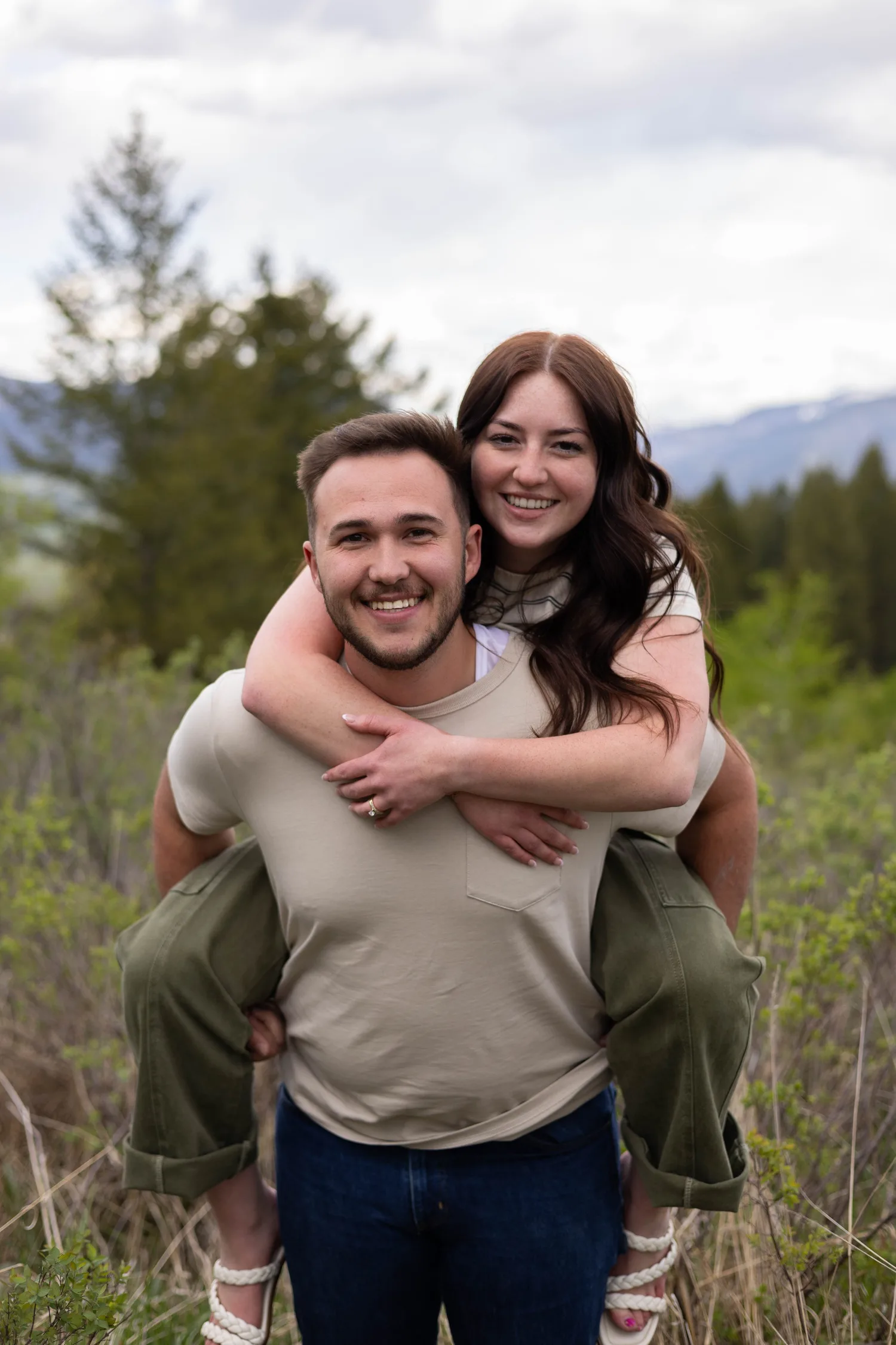 Woman riding piggyback on her fiancé, both smiling, in a spring meadow