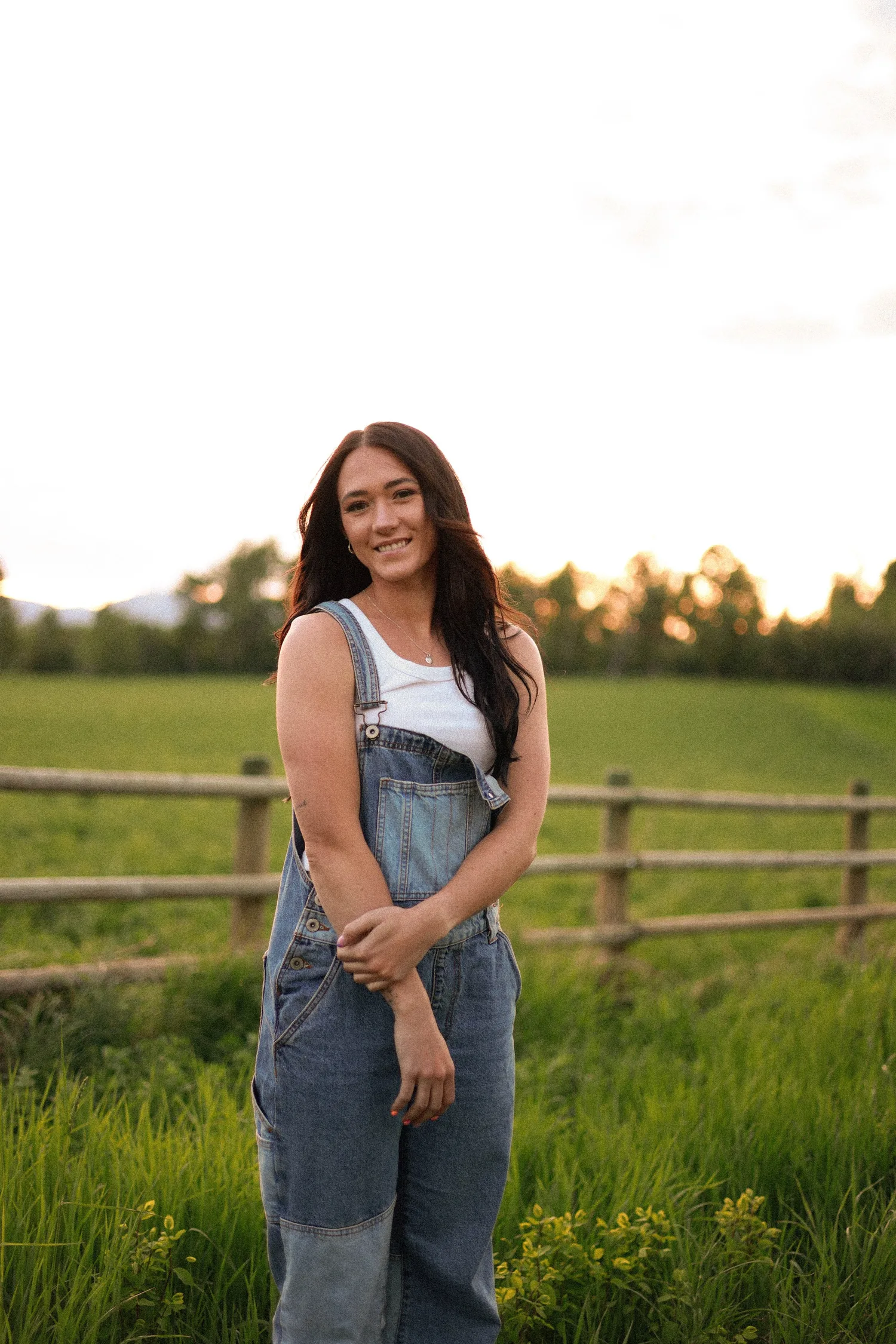 Senior portrait of a young woman in denim overalls by a wooden fence at golden hour